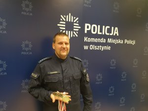 &quot;Bój o Niepodległość&quot; - zdjęcie na tle fototapety KMP w Olsztynie nadkom. Tomasza Okorskiego, który w prawy ręku trzyma trzy złote medale zdobyte w Prabutach podczas zawodów strzeleckich.