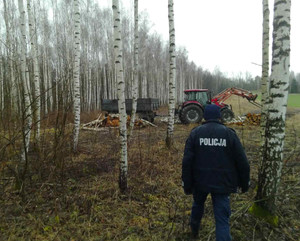 Zdjęcie przedstawia policjanta a w tle ciągnik rolniczy i drewno.